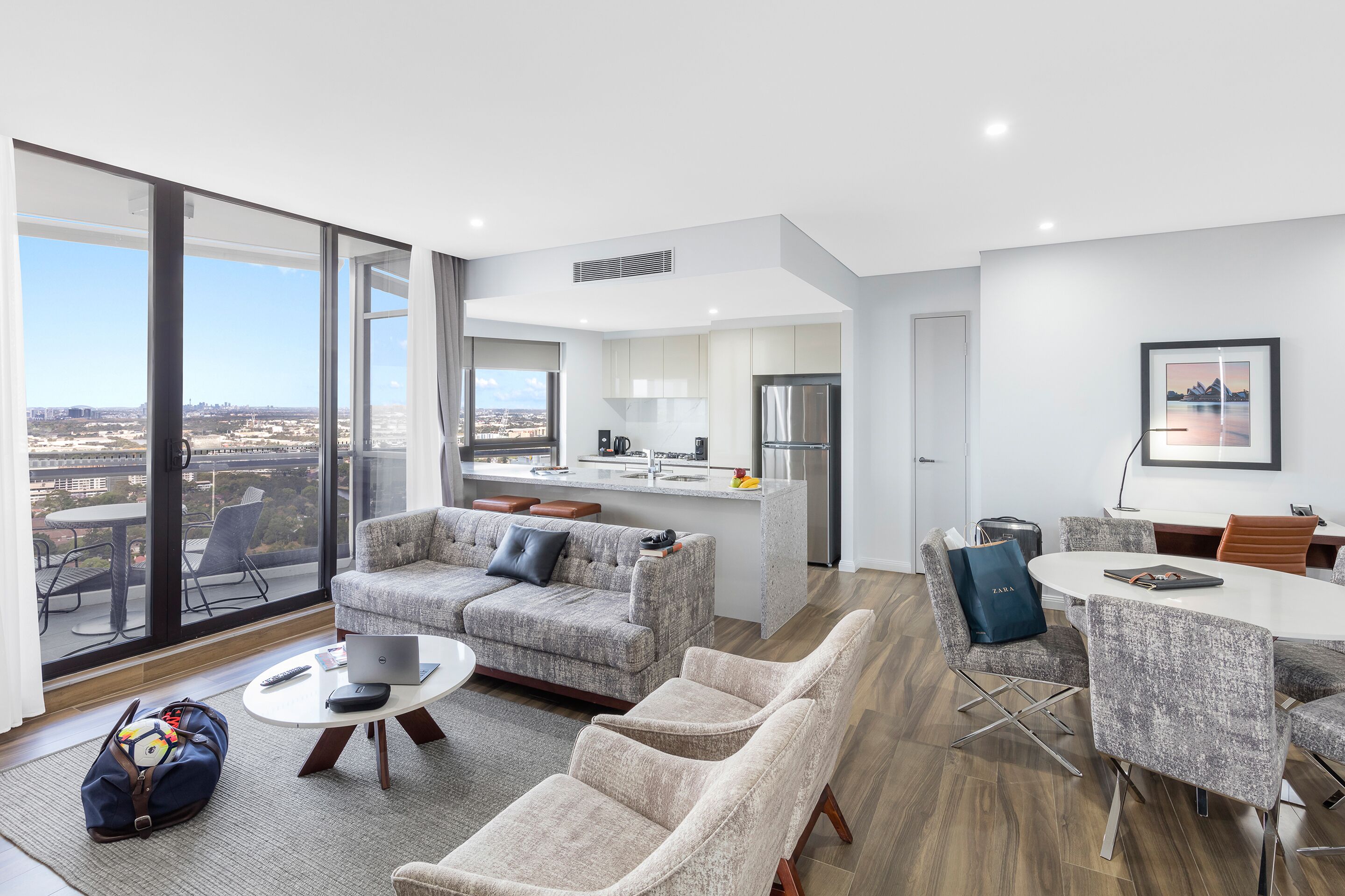 A modern living room with a sofa, armchairs, a coffee table, and a dining area with a table and chairs. A large window offers a view of the city.