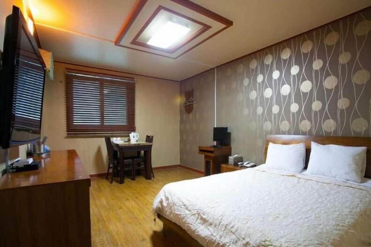 Habitación de hotel con cama, escritorio y silla, una mesita, televisor y ventana con persianas.