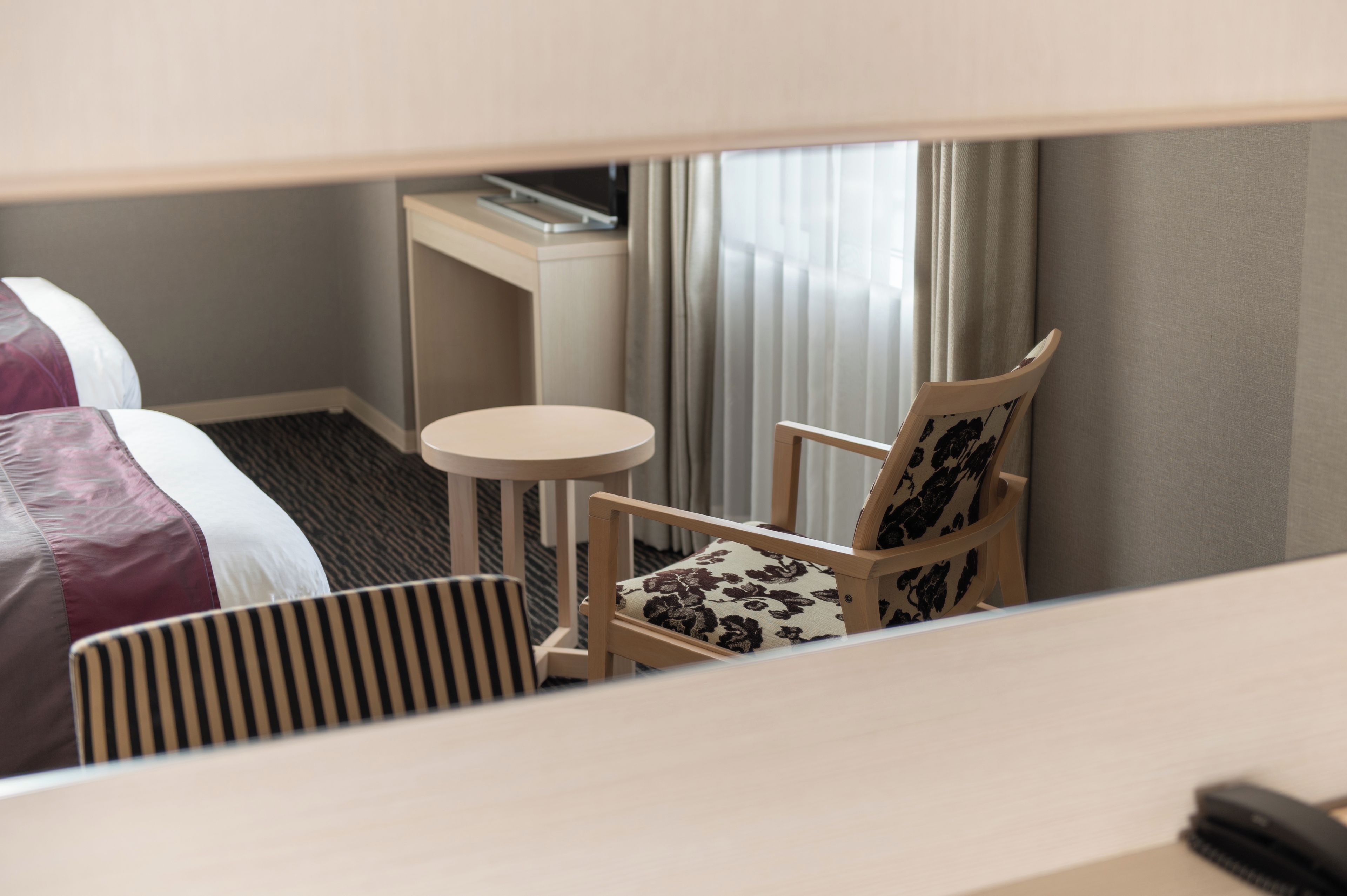 A hotel room with a bed, a small round table, a chair with a floral cushion, and a desk with a lamp.