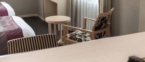 A hotel room with a bed, a small round table, a chair with a floral cushion, and a desk with a lamp.