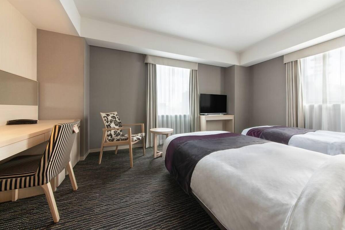 A hotel room with a desk, two beds, a TV, and a chair.