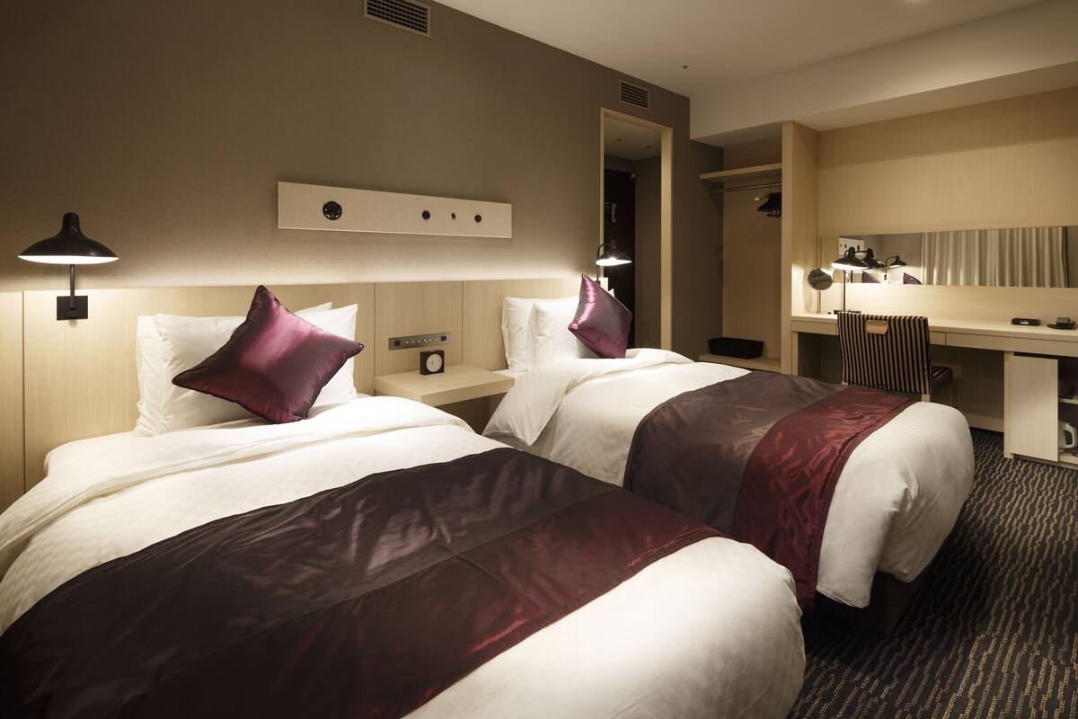 A hotel room with two beds, a desk, a chair, a mirror, and a lamp.