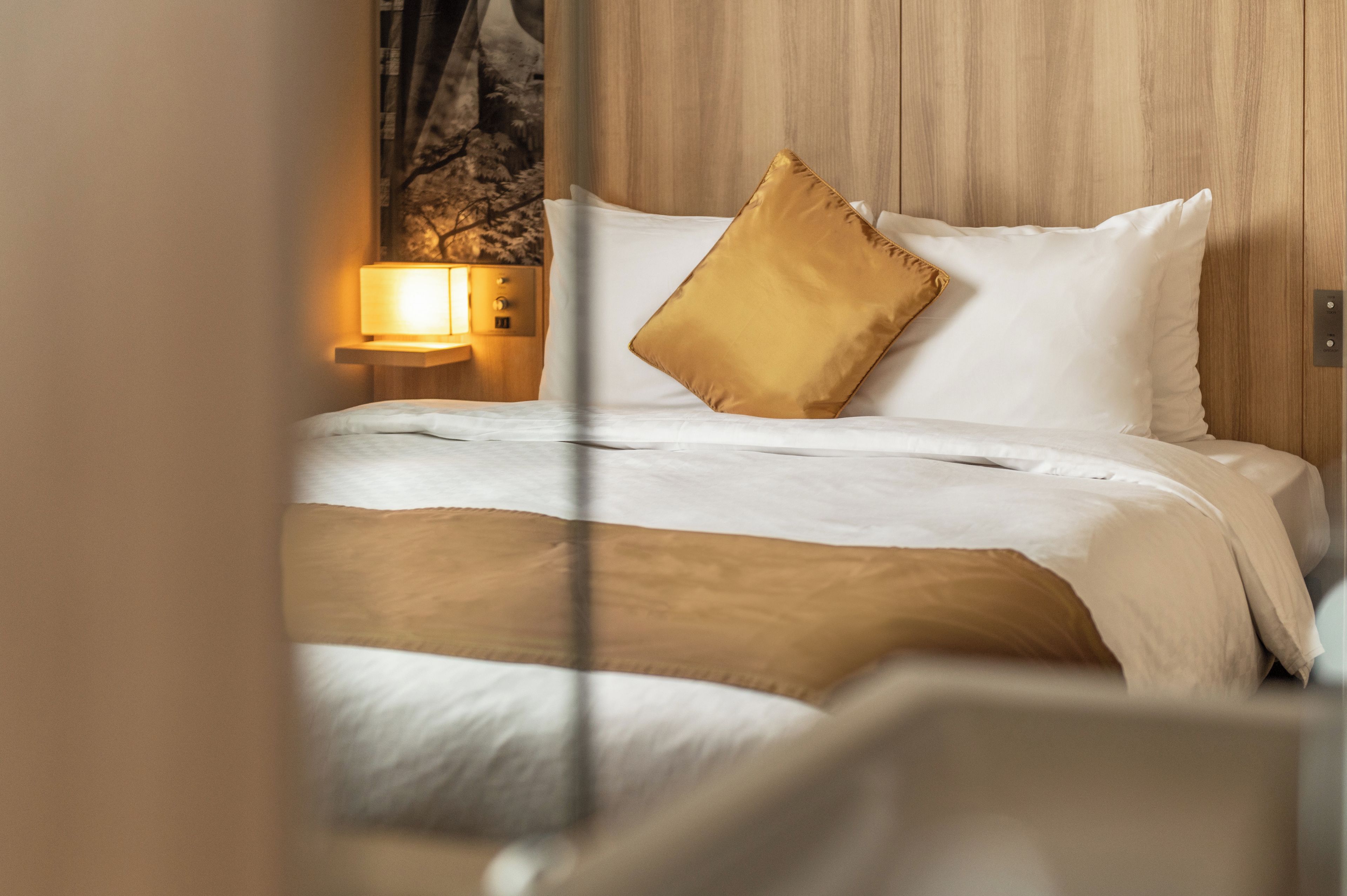 A neatly made bed with white linens and a single gold-colored pillow, a bedside lamp, and a wooden headboard.