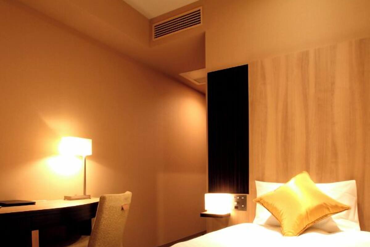 A hotel room with a bed, a desk, a chair, and a lamp.