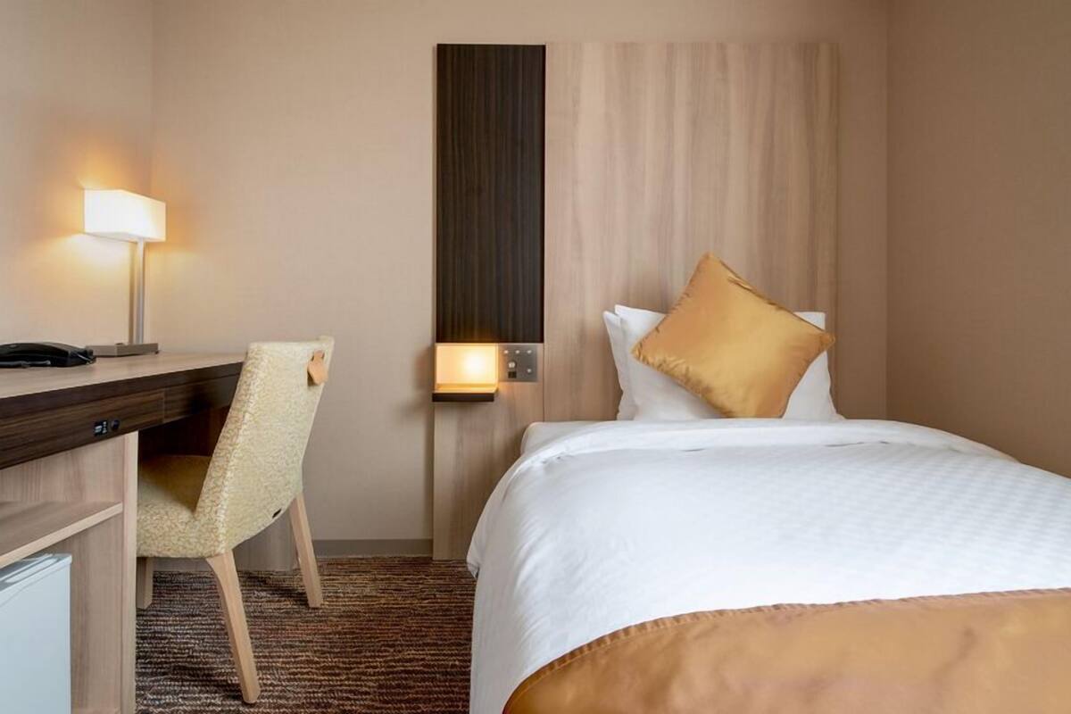 A hotel room with a bed, a desk, a chair, a lamp, and a wall-mounted control panel.
