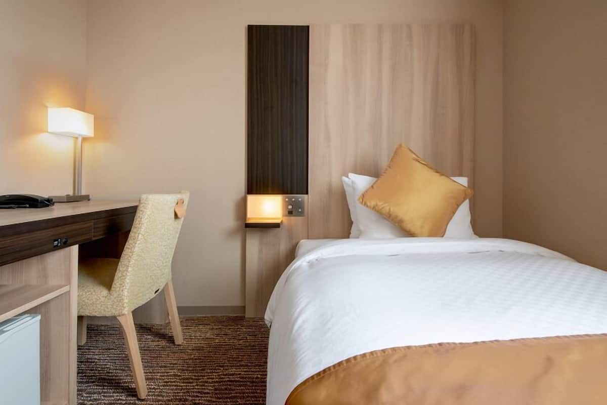 A hotel room with a bed, a desk, a chair, a lamp, and a wall-mounted control panel.