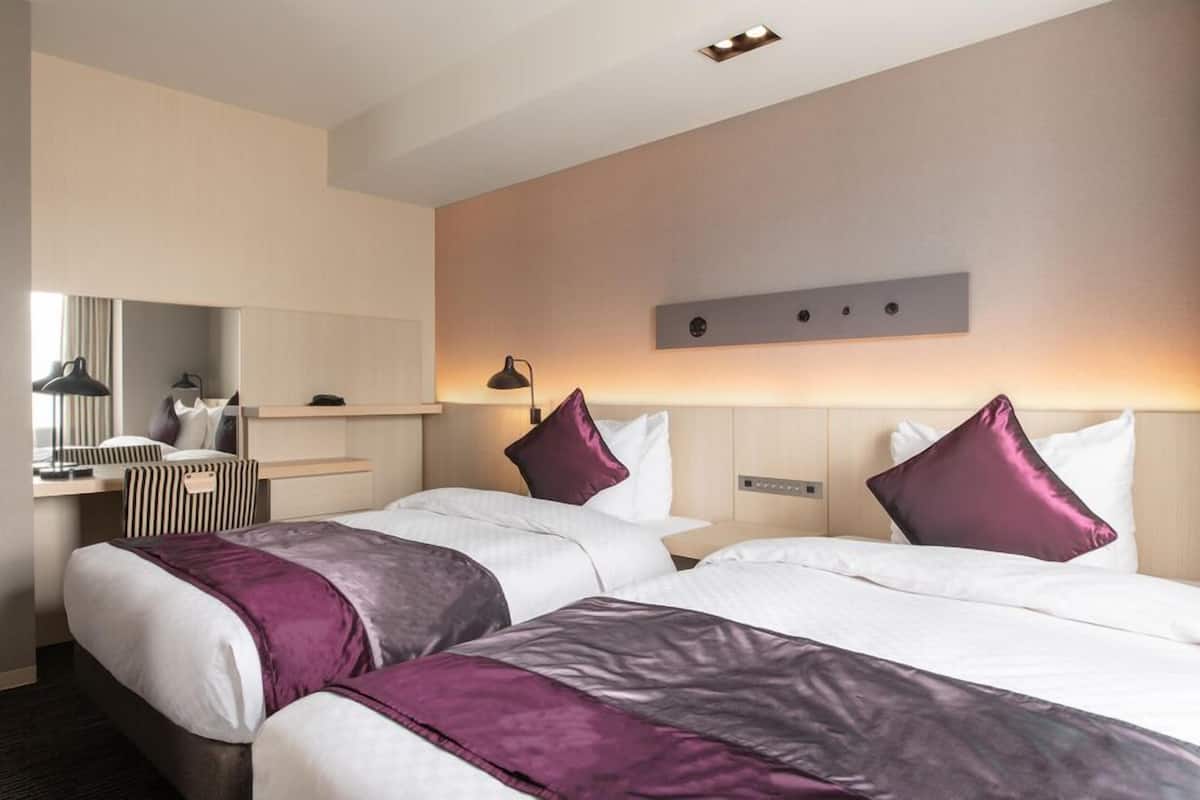 A hotel room with two beds, a desk, a mirror, and a wall-mounted light fixture.