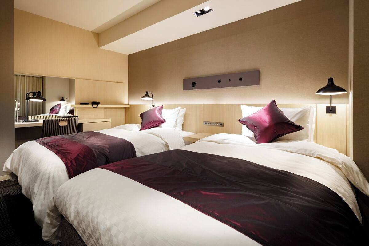 A hotel room with two beds, a desk, a mirror, and a wall-mounted light fixture.