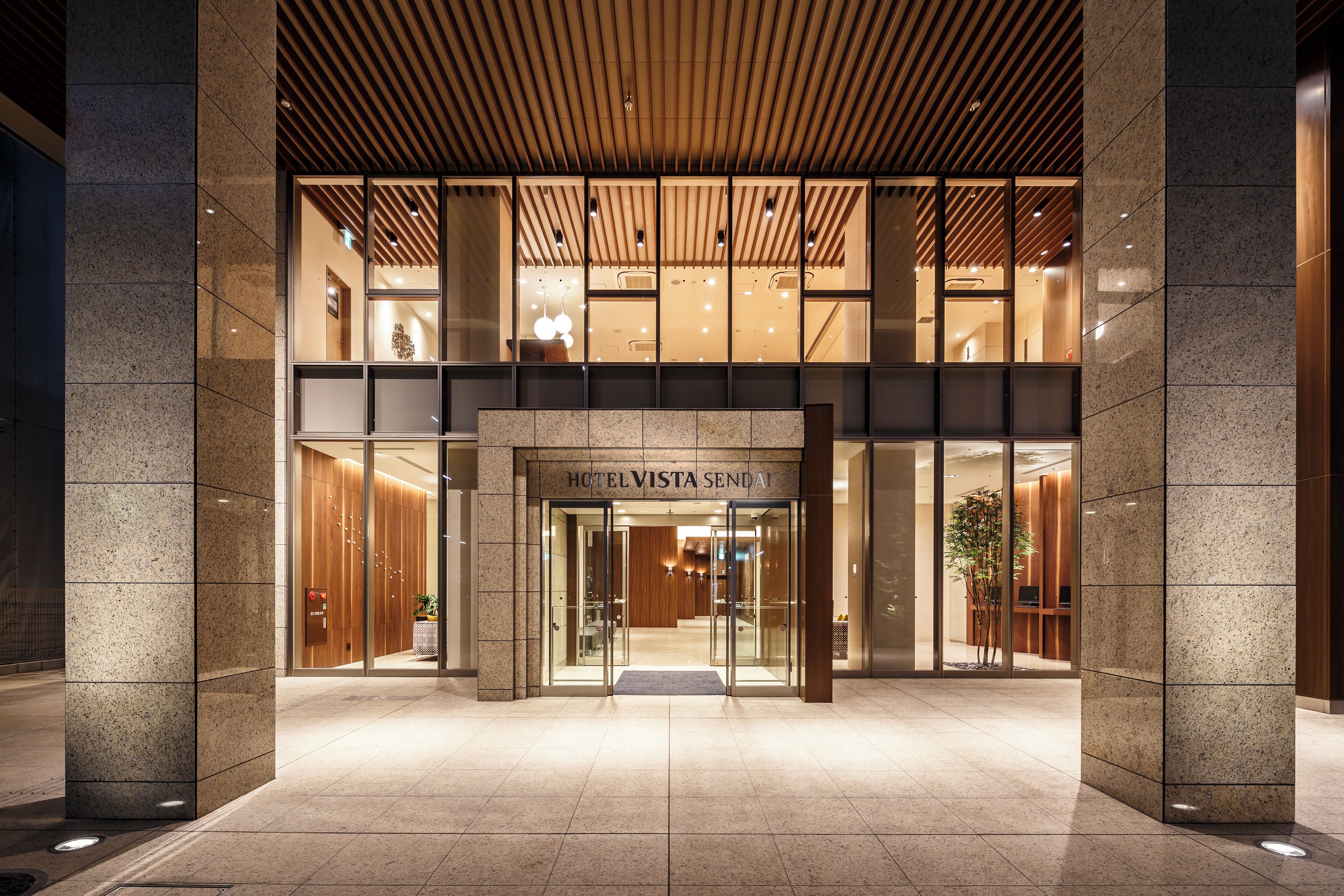 A hotel entrance with a modern design, featuring a glass facade, a wooden ceiling, and a sign that reads "HOTEL VISTA SENDA".