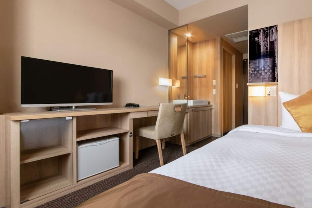 A hotel room with a bed, a desk with a TV, a chair, a small fridge, and a mirror.