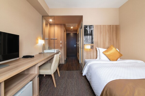 A hotel room with a bed, a desk with a TV, a chair, a small bathroom, and a door leading to another room.