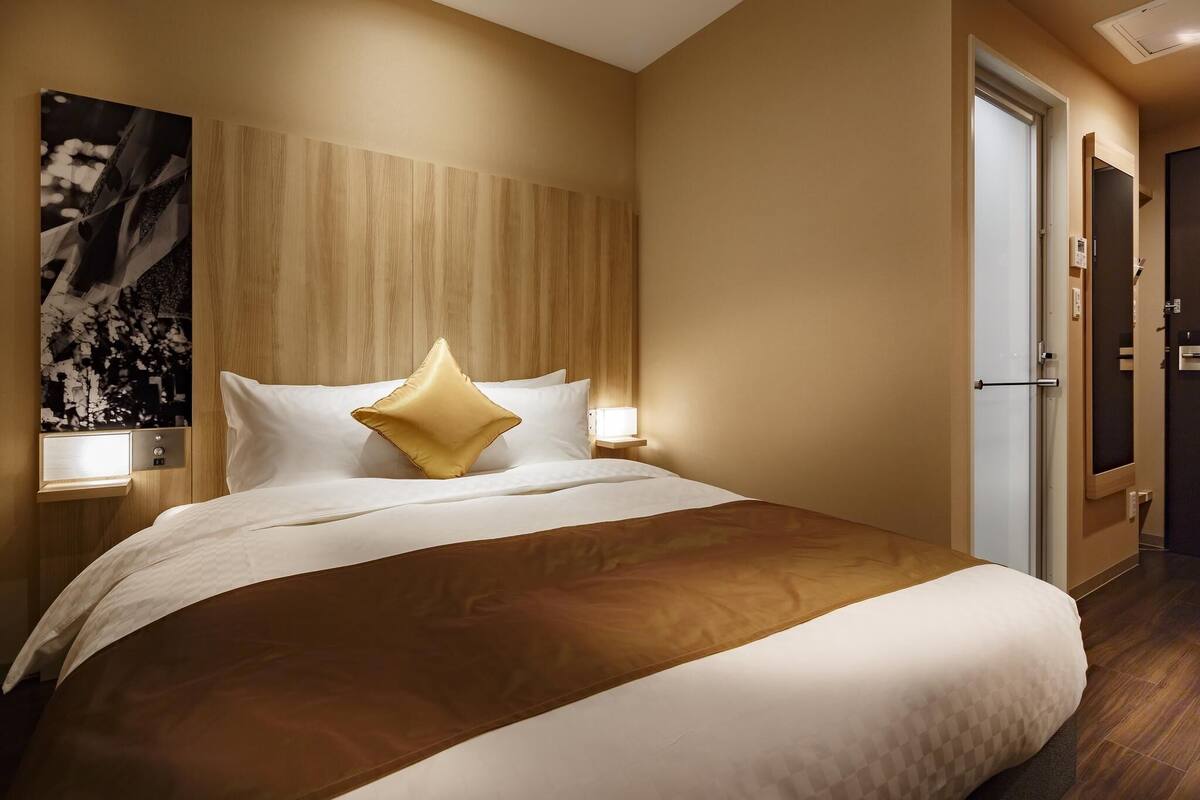 A hotel room with a large bed, a wooden headboard, a nightstand with a lamp, and a door to the right.
