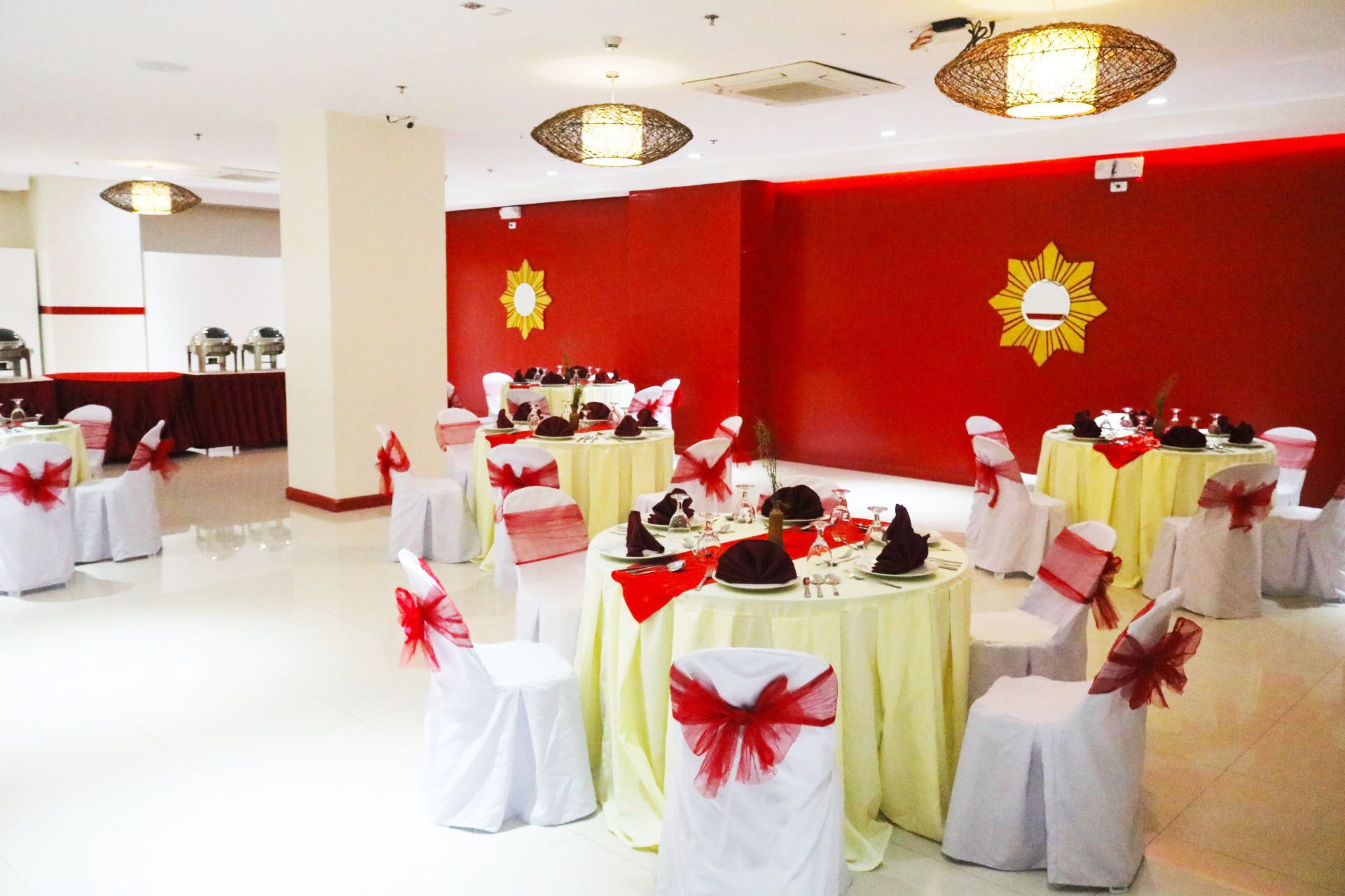 Un salón de banquetes con mesas redondas, sillas blancas con cintas rojas y paredes rojas decoradas con motivos de rayos de sol.