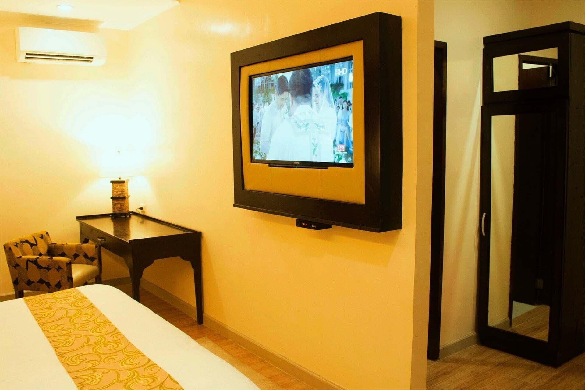 A hotel room with a bed, a small bedside table, a TV mounted on the wall, and a door.
