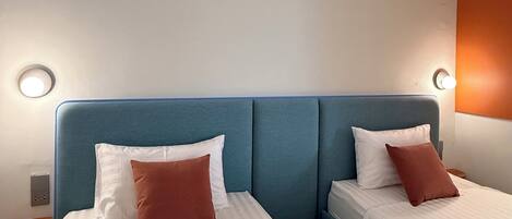 Quarto de hotel com duas camas, cada uma com roupa de cama branca e uma almofada marrom. As cabeceiras são verde-água, e há uma pequena mesa de cabeceira de madeira com uma luminária entre as camas.