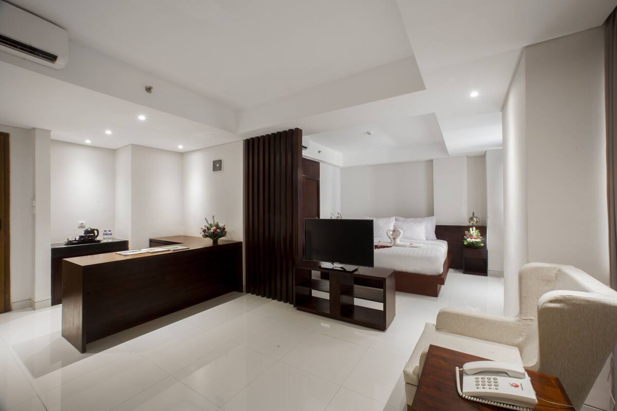 A modern hotel room with a reception desk, a flat-screen TV, a bed, a sofa, and a telephone.