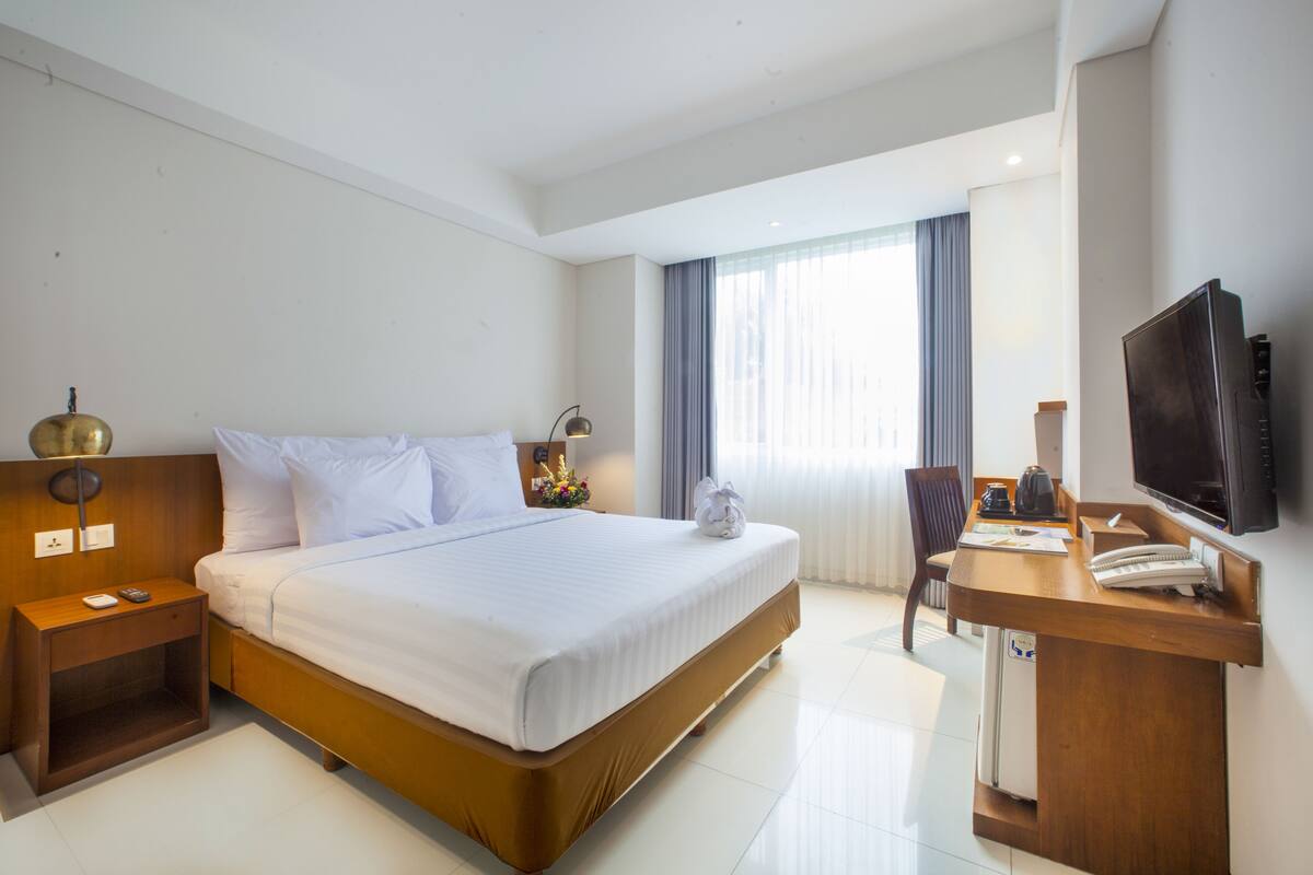 A hotel room with a large bed, bedside tables, a desk with a chair, a television, and a window with curtains.