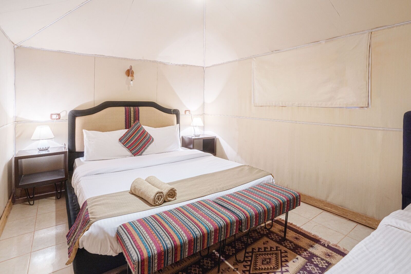 A hotel room with a bed, two bedside tables, a lamp, a window, and a patterned rug.