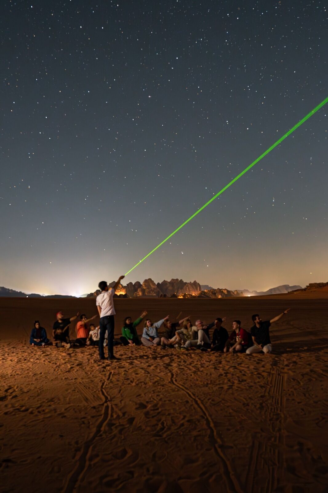 A group of people sitting on a sandy beach under a starry sky, with a green laser beam in the sky.