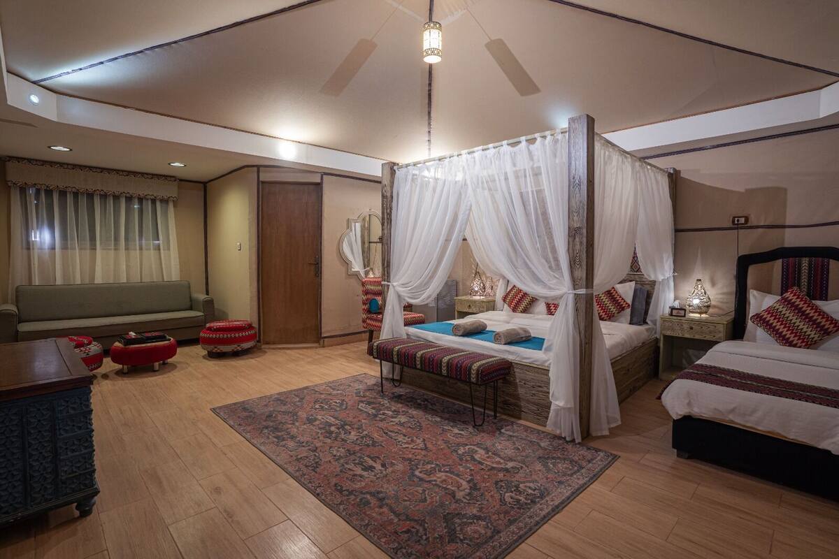 A spacious bedroom with a four-poster canopy bed, a sofa, a small table, and a nightstand with a lamp.