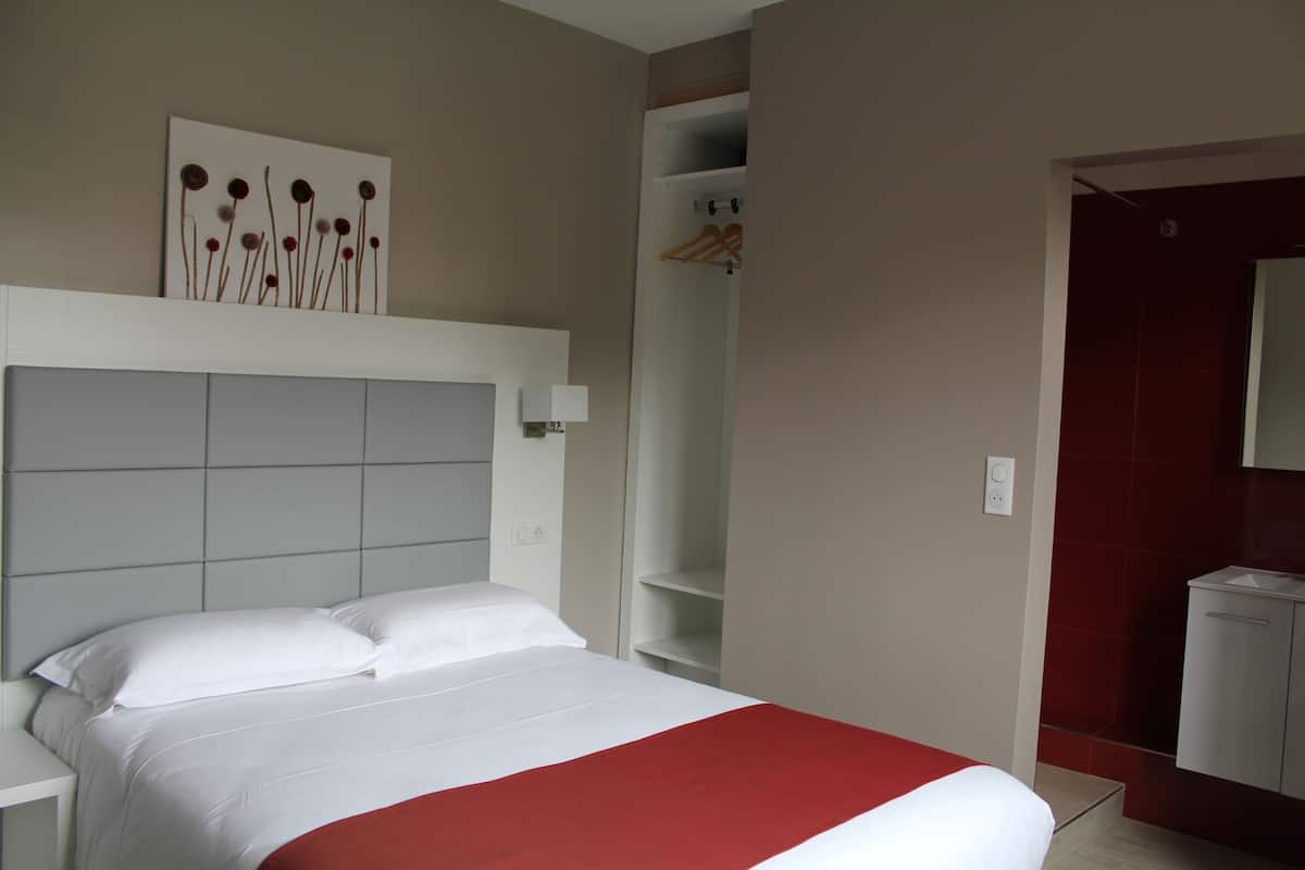 Une chambre d’hôtel avec un lit, une tête de lit blanche, un élément décoratif rouge sur le lit, une table de chevet blanche, une lampe fixée au mur, une étagère blanche, une porte rouge et une salle de bain blanche avec un miroir.