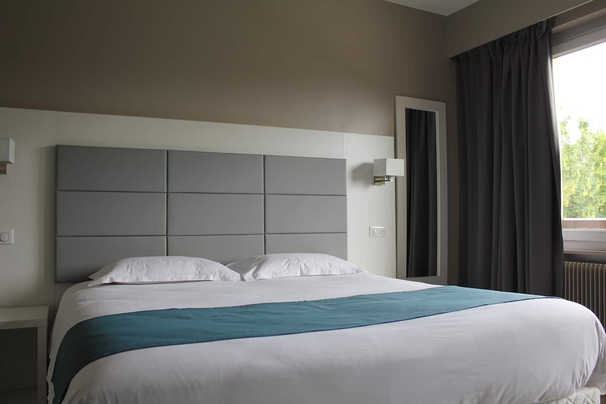 Une chambre d’hôtel avec un grand lit, une tête de lit dotée d’un interrupteur, une table de chevet et une fenêtre avec des rideaux.