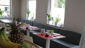 Comedor con una mesa larga, asientos tipo banca con cojines grises y plantas en macetas en los alféizares de las ventanas.