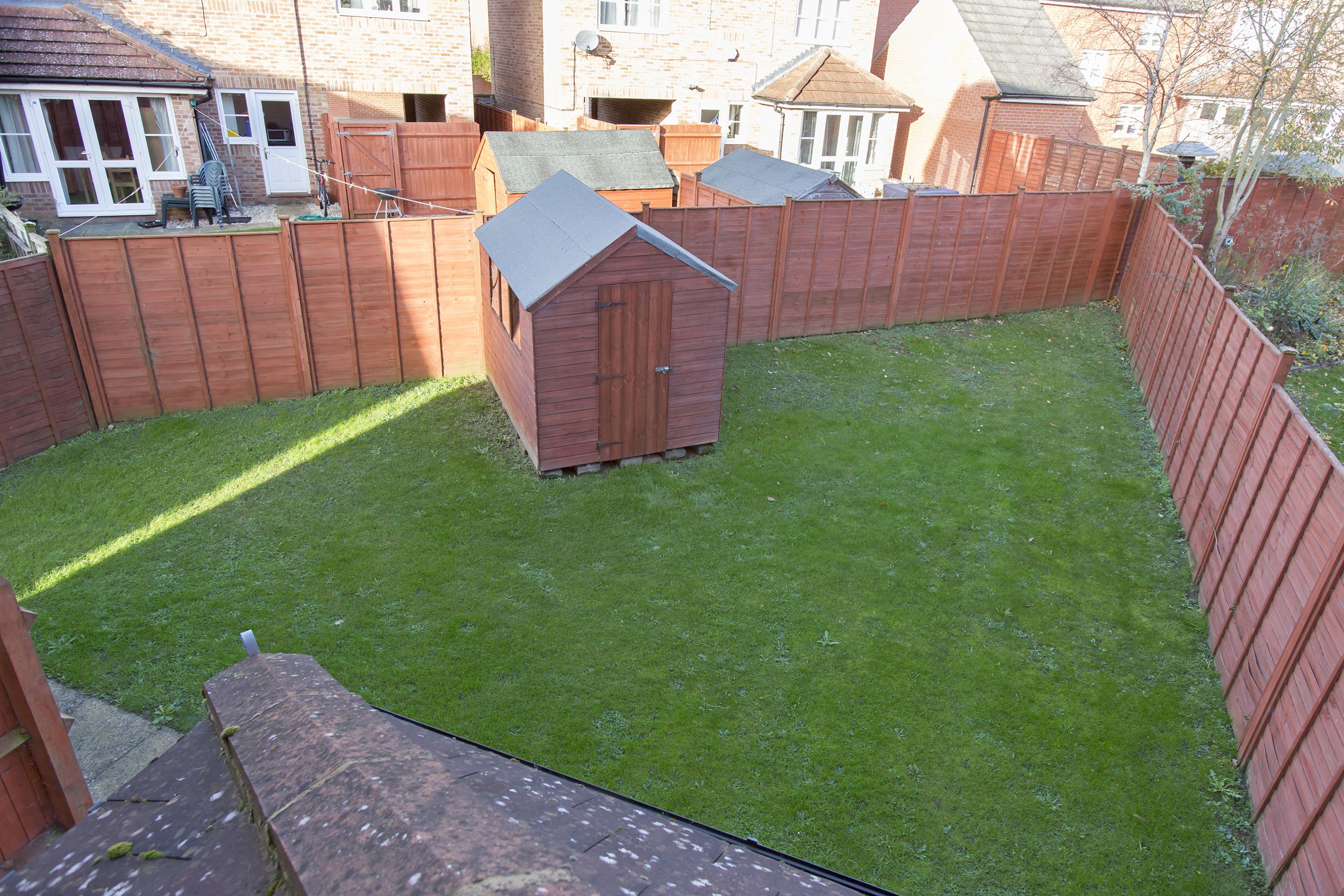 A fenced backyard with a small wooden shed, a well-maintained lawn, and a brick house in the background.