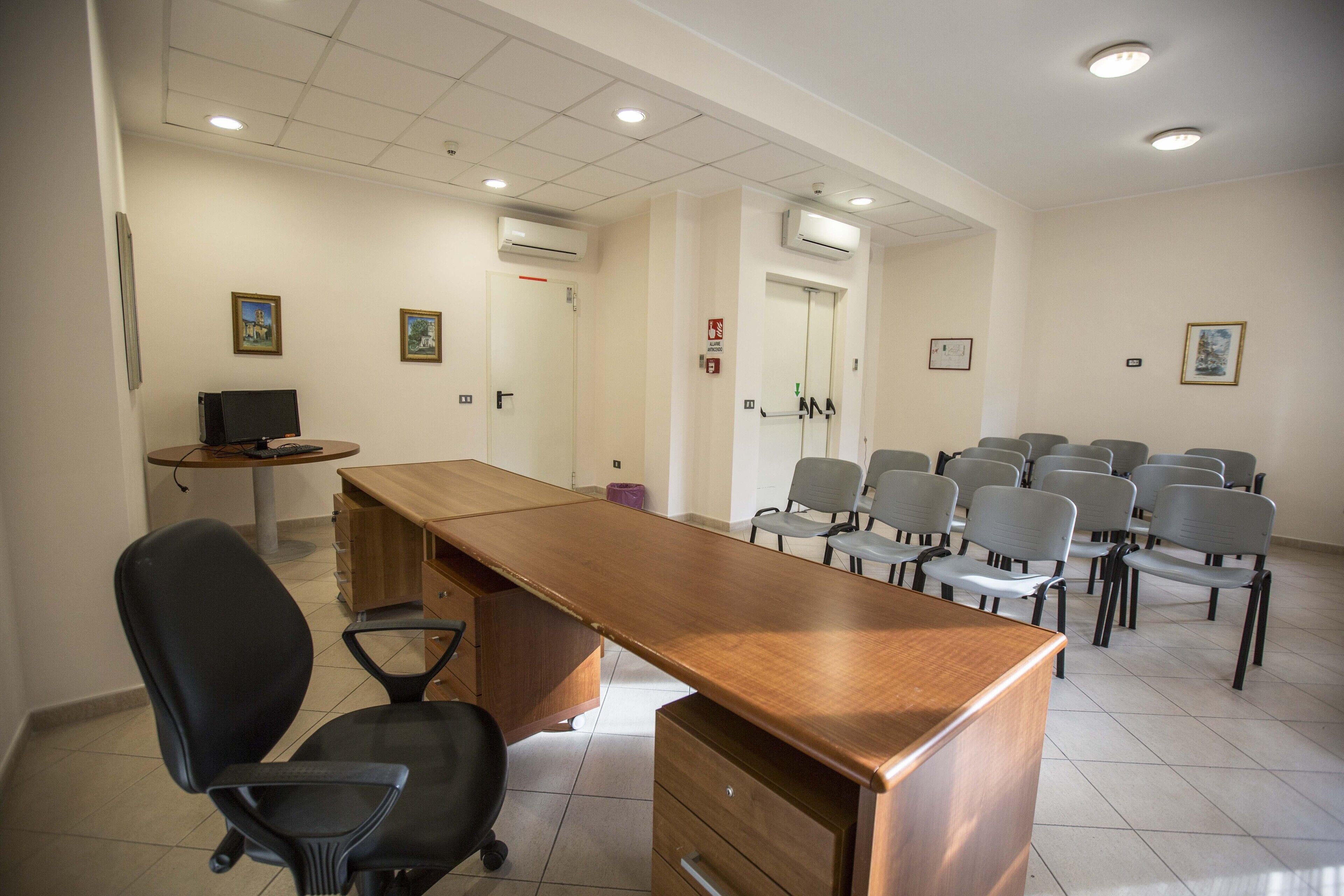 Un salón de conferencias con una larga mesa de madera, varias sillas, un escritorio con una computadora y unidades de aire acondicionado en las paredes.