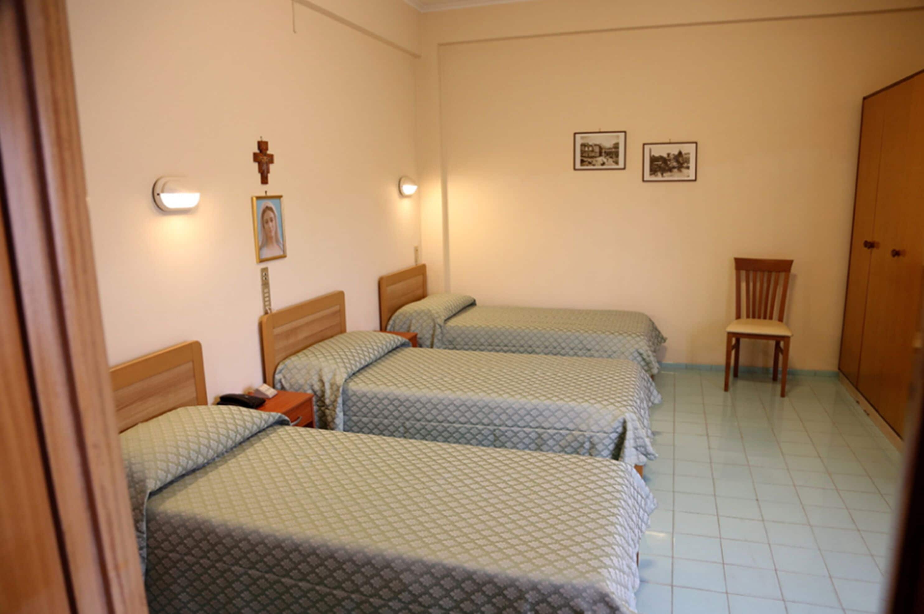 Une pièce avec trois lits, une chaise en bois et une croix accrochée au mur.