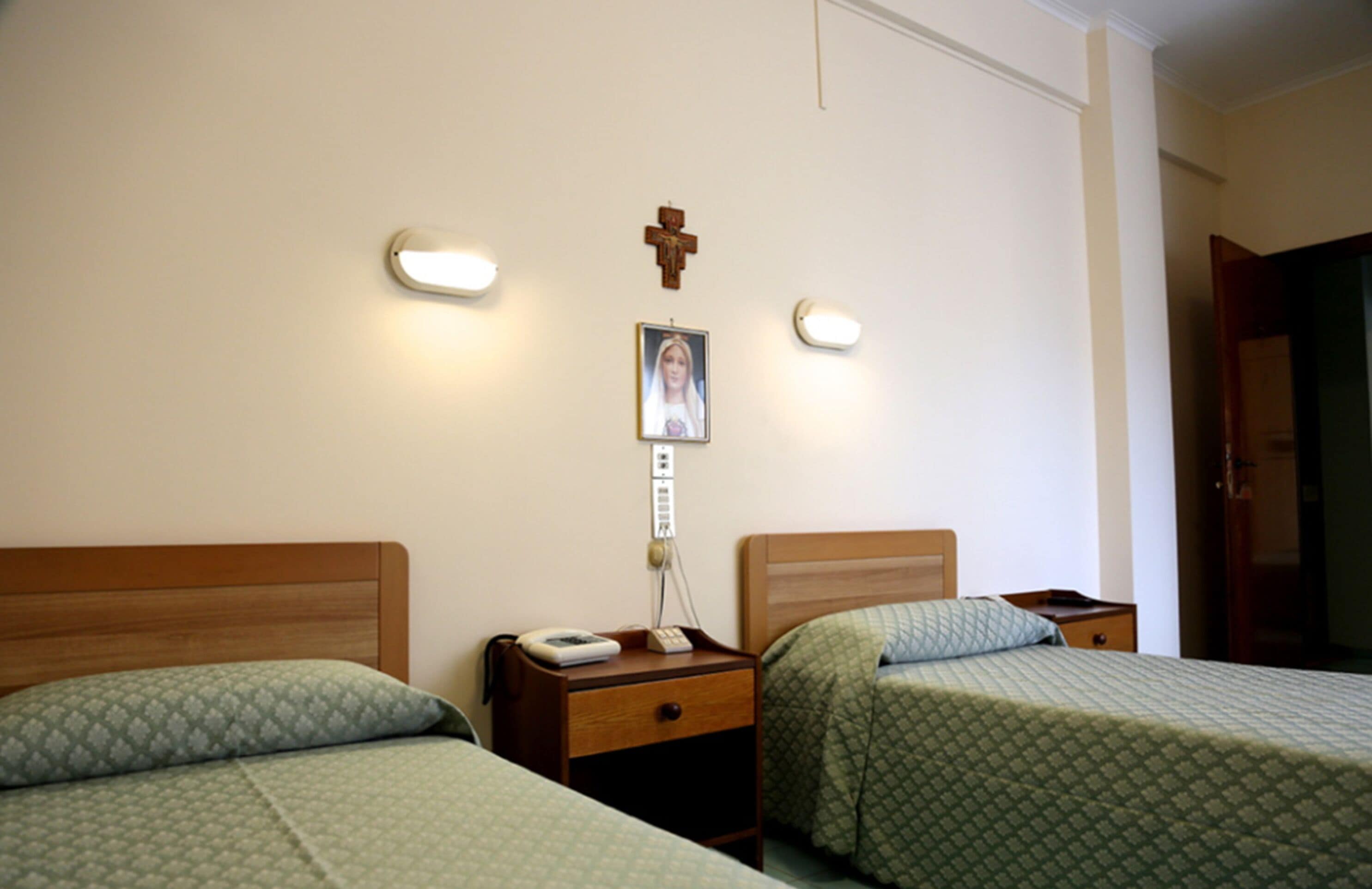 Habitación de hotel con dos camas individuales, mesitas de noche, una cruz colgada en la pared y un cuadro enmarcado.