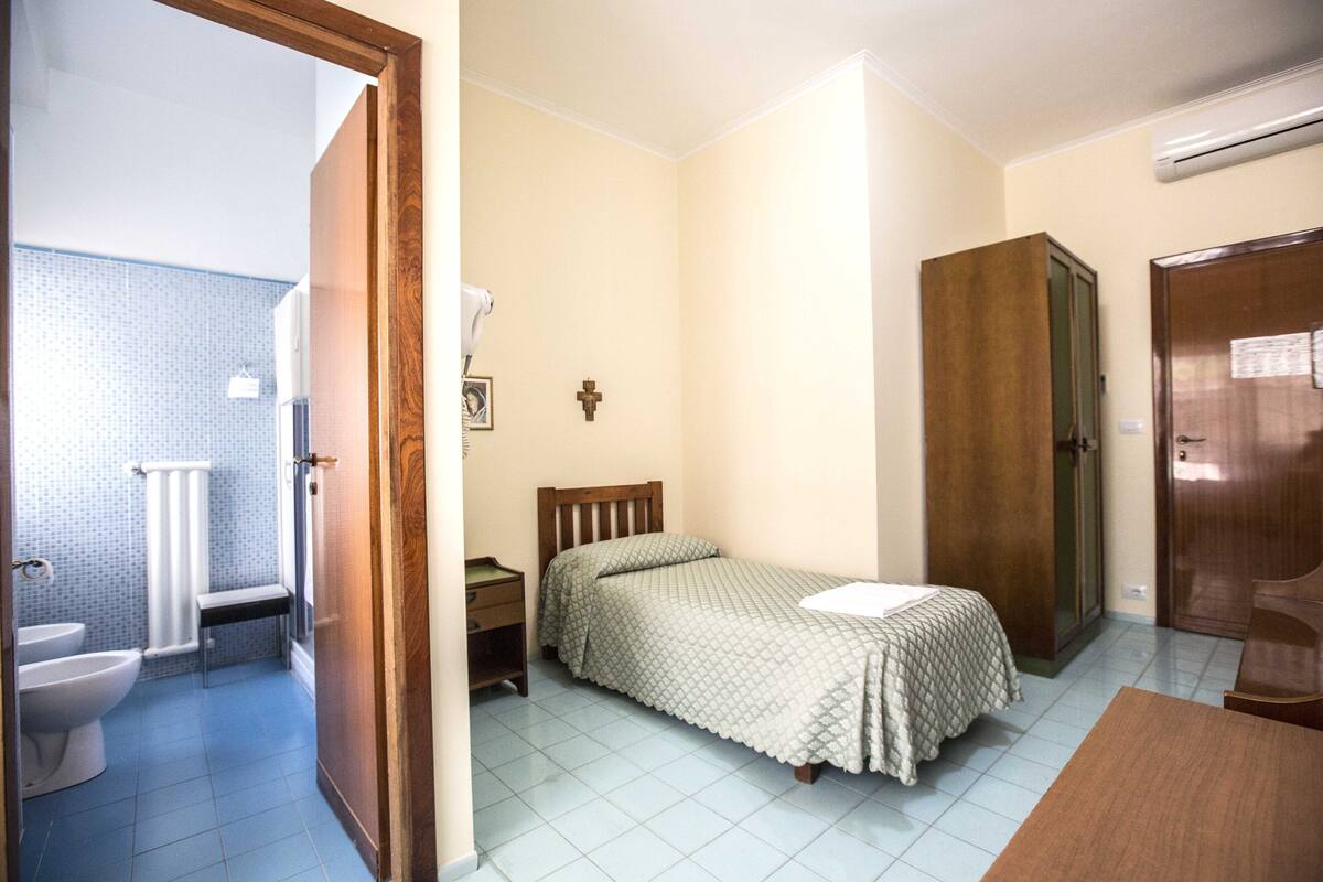 Een slaapkamer met een enkel bed, een houten garderobe, een nachtkastje en een badkamer die door een open deur zichtbaar is.