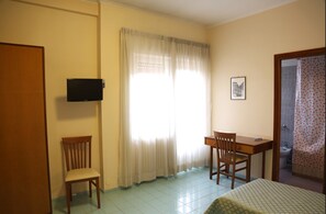 Une pièce comprenant un lit, deux chaises en bois, un bureau, un téléviseur fixé au mur et une fenêtre avec des rideaux.