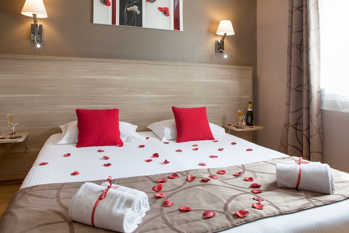 A hotel room with a bed, two red pillows, a nightstand with a bottle and glass, and a wall-mounted lamp.