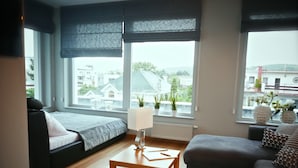 Ein modernes Wohnzimmer mit einer Couch, einem kleinen Tisch und großen Fenstern mit Blick auf Gebäude und Grünflächen.