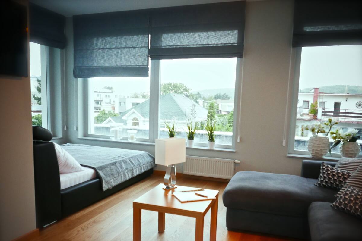 A modern living room with a sofa, a small table, and large windows offering a view of buildings and greenery.