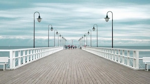 Eine hölzerne Pier mit weißer Reling, gleichmäßig verteilte Straßenlaternen und ein Blick aufs Meer in der Ferne.