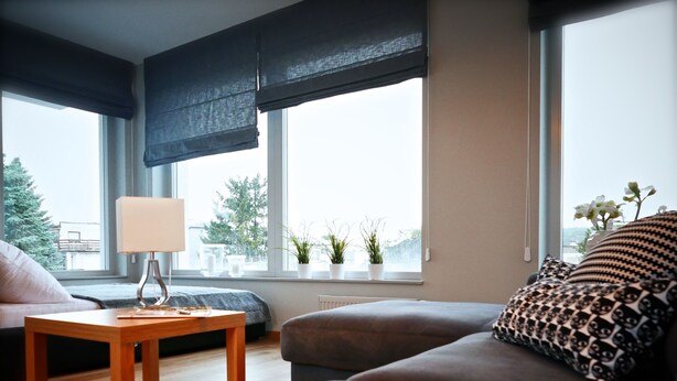 Ein modernes Wohnzimmer mit einer Couch, einem kleinen Holztisch und einer Lampe. Große Fenster mit Jalousien bieten Blick auf Bäume und Gebäude.