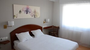 A neatly made bed with white linens, a wooden headboard, a desk with a chair, and a wall-mounted lamp. - Hôtel le Petit Trianon (Saint-Brevin-les-Pins)