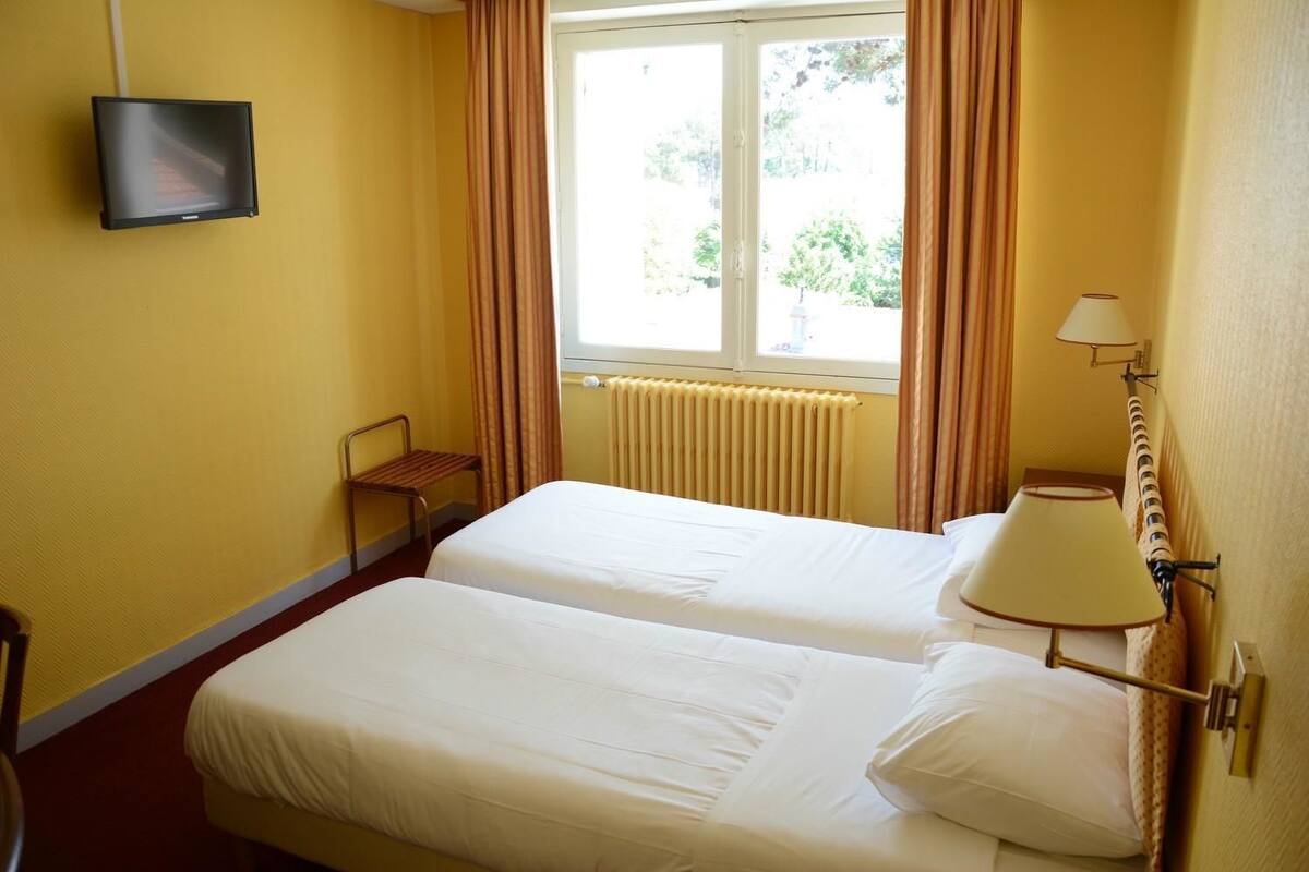 Ein Hotelzimmer mit zwei Betten, einem Fernseher, einem Fenster mit Vorhängen, einem Heizkörper und einer Lampe.