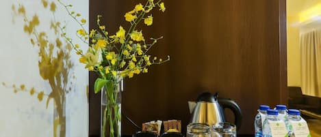 A hotel room with a wooden sideboard, a vase with yellow flowers, a kettle, and bottled water.