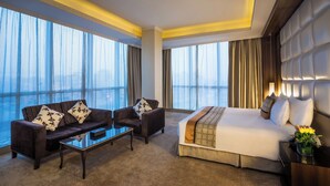 Ein Hotelzimmer mit einem großen Bett, einer Couch, einem Couchtisch und Blick auf die Stadt durch große Fenster.