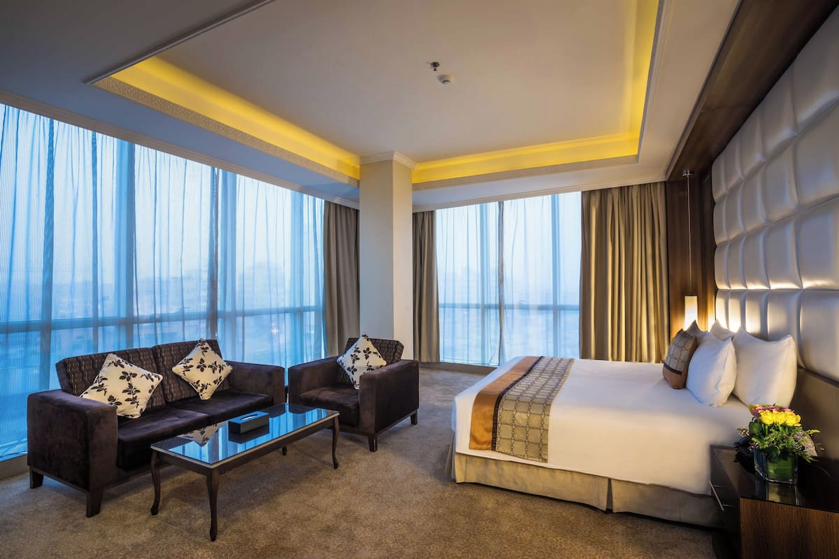 Una habitación de hotel con una cama grande, un sofá, una mesa de centro y vistas a la ciudad a través de amplias ventanas.