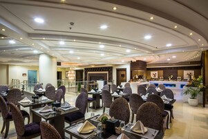 A spacious hotel dining area with multiple tables and chairs, a reception desk, and a modern ceiling design.