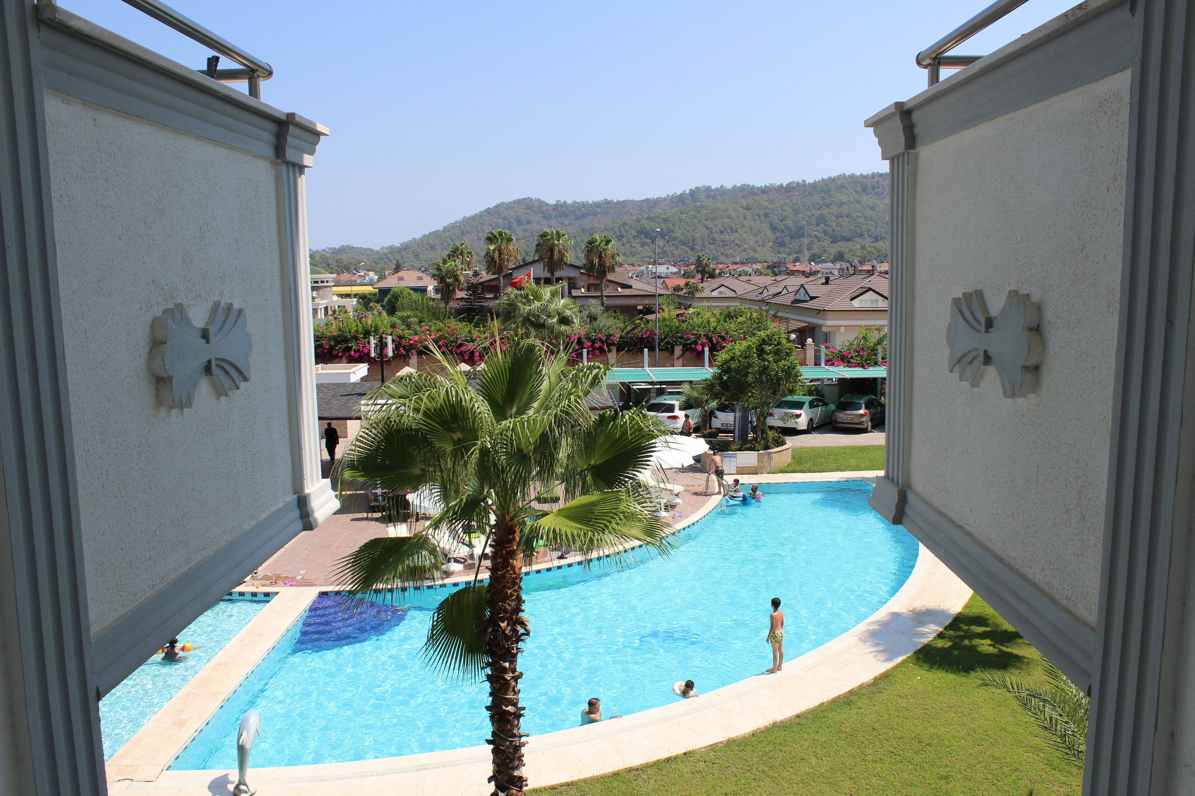 Vista di una piscina con palme, circondata da un prato curato e edifici sullo sfondo.