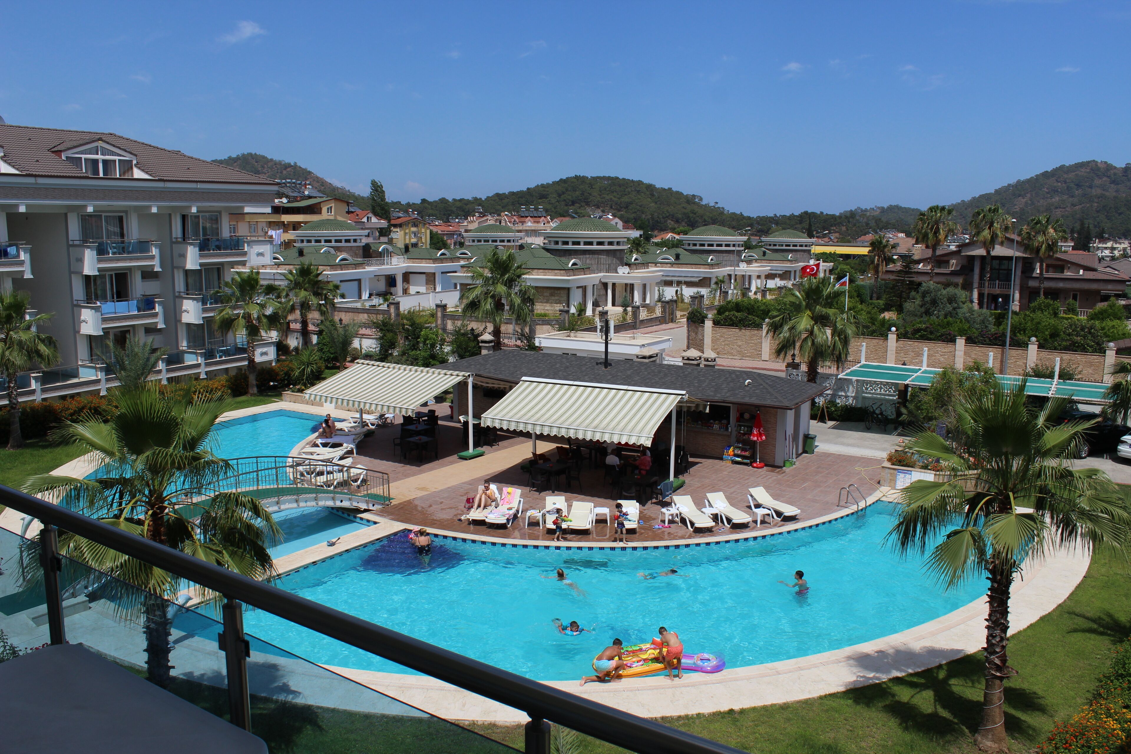 A resort with a large swimming pool, multiple lounge areas, and a view of surrounding buildings and hills.