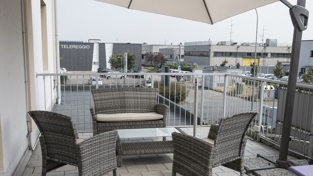 A balcony with wicker furniture, a glass table, and a view of a parking lot and buildings. - Hotel Corte Business (Cavriago)