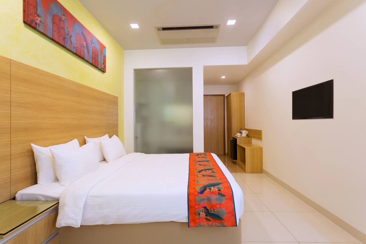 A hotel room with a bed, a nightstand, a wooden headboard, a wall-mounted TV, and a painting above the bed.