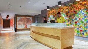 Un comptoir d’accueil agrémenté d’un mur décoré d’une mosaïque colorée, un bureau d’accueil en bois et un couloir doté d’un plancher en bois et d’une fresque représentant un arbre.