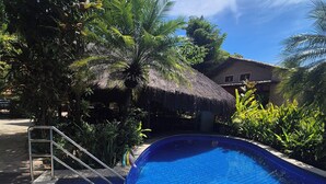 Una piscina con borde de azulejos, rodeada de exuberante follaje y una estructura de techo de paja.